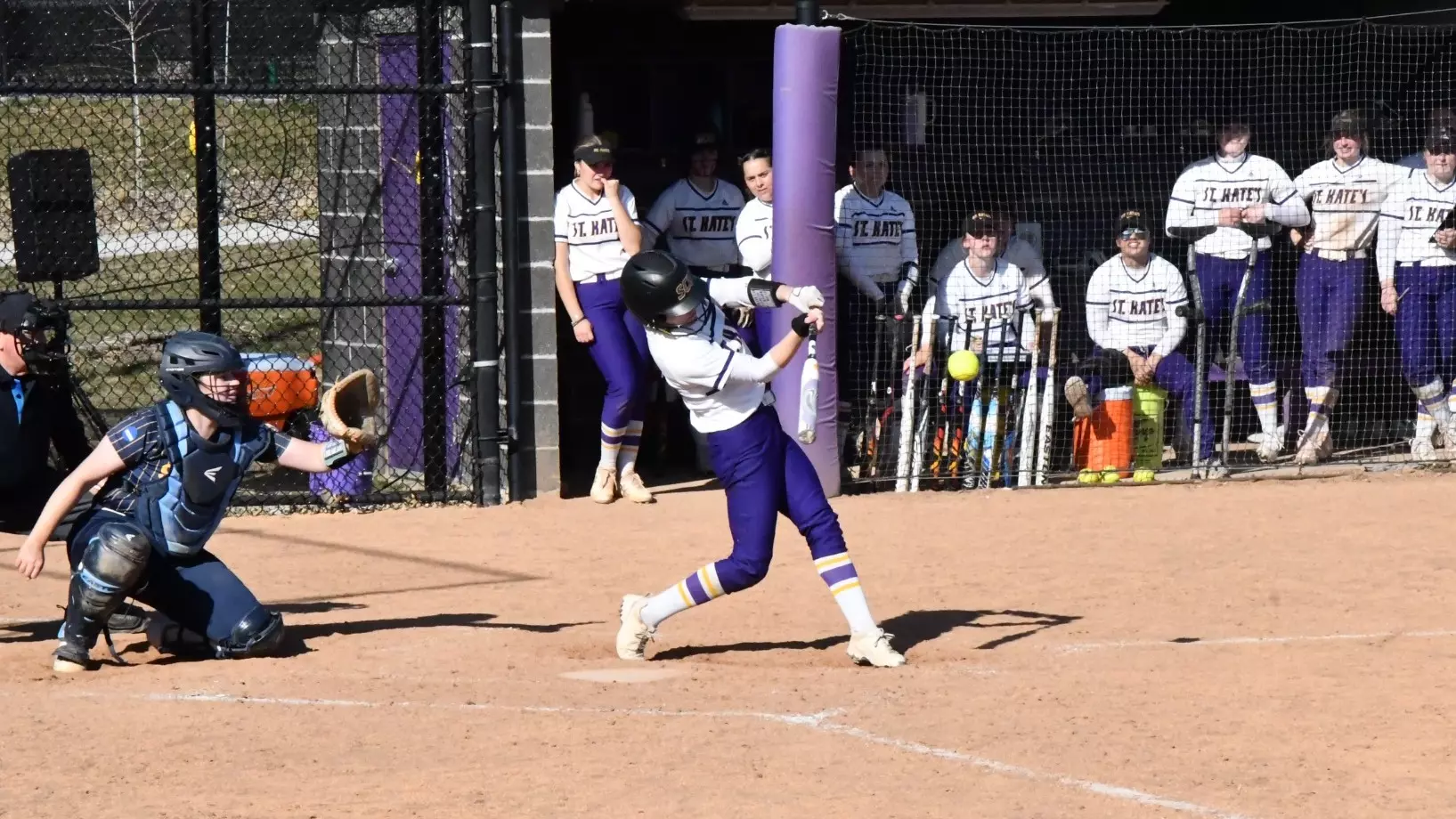 Lynae Doty making contact with softball at home as team watches on from dugout